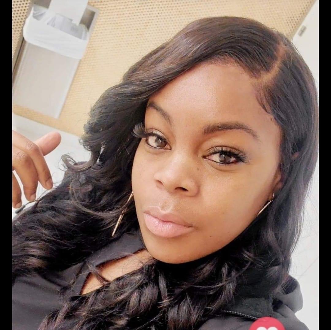 Close-up portrait of a woman with wavy dark hair, makeup, and hoop earrings, taking a selfie in an indoor setting.