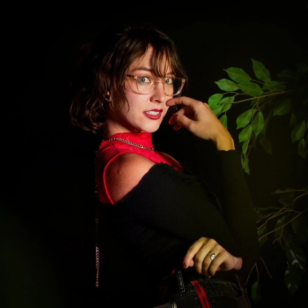 A woman with short brown hair and glasses, wearing a black top with red accents, posing against a black background with green foliage.