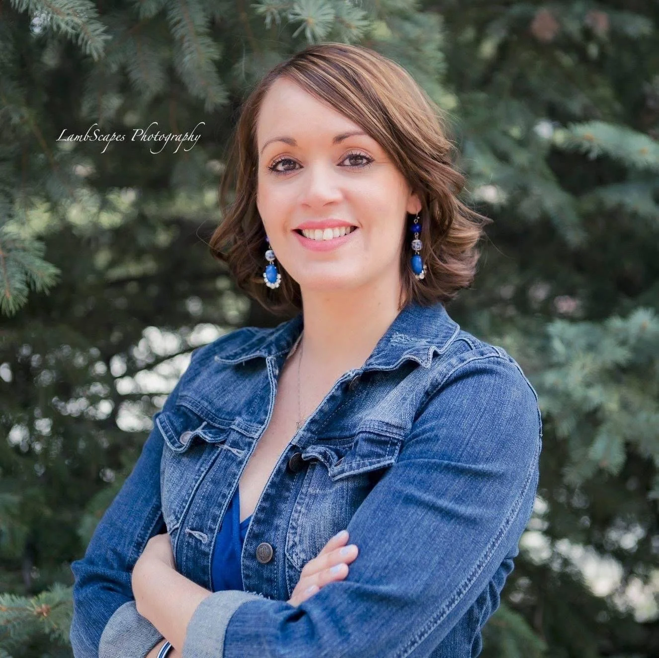 A woman with shoulder-length brown hair and blue earrings smiling with arms crossed, standing outdoors in front of a pine tree.