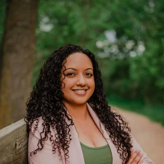 A woman with curly dark hair smiling outside, leaning against a wooden railing near a tree and green foliage.
