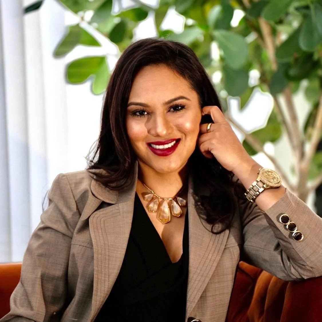 A woman with dark hair and a warm smile, wearing a beige blazer, black top, and gold jewelry, sitting indoors with green plants in the background.