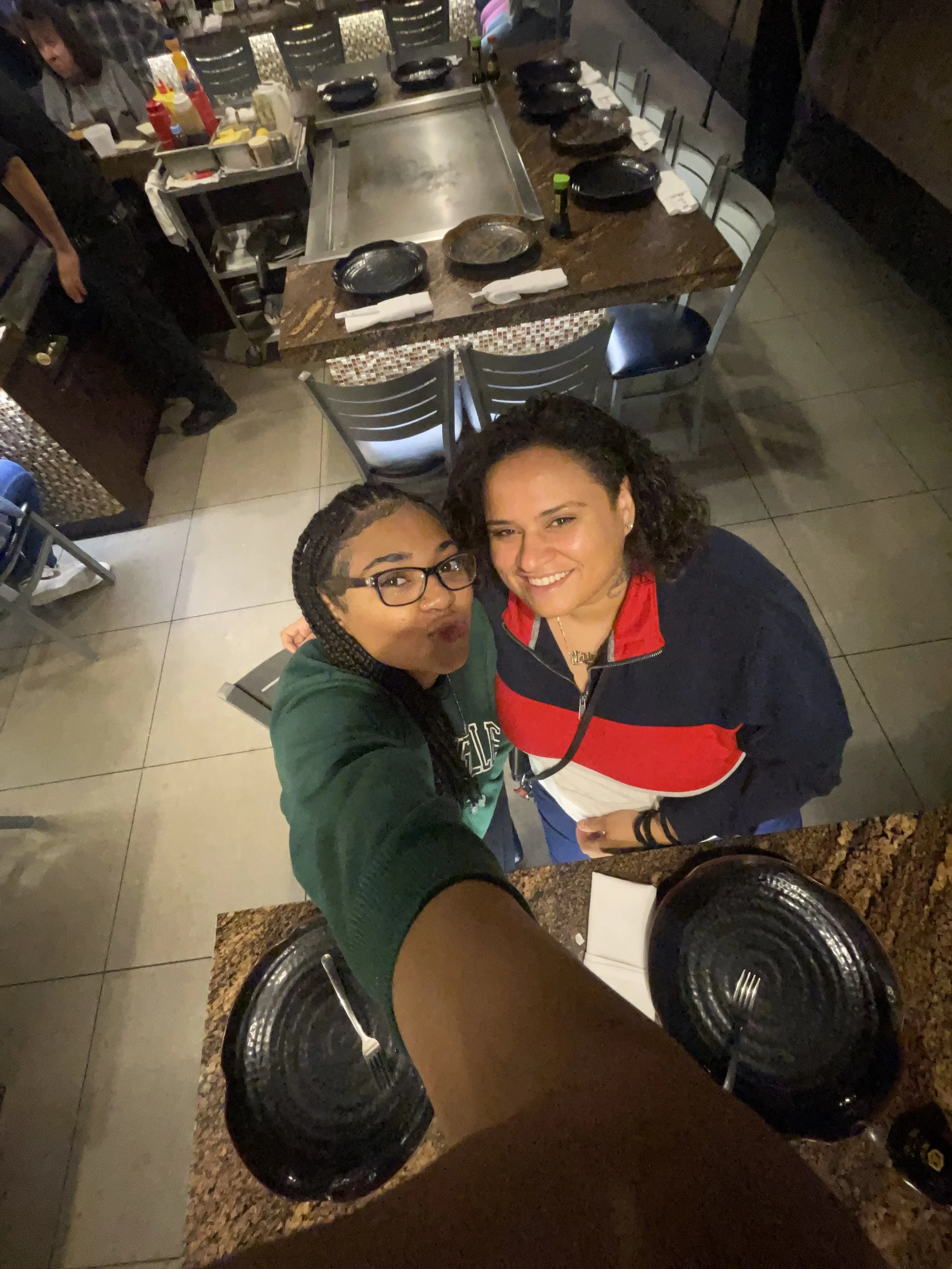 Two women smiling and taking a selfie at a restaurant, viewed from above. The woman on the left has glasses and braided hair, and the woman on the right has curly hair and is wearing a red and black jacket. The table below has black plates, napkins, and utensils.