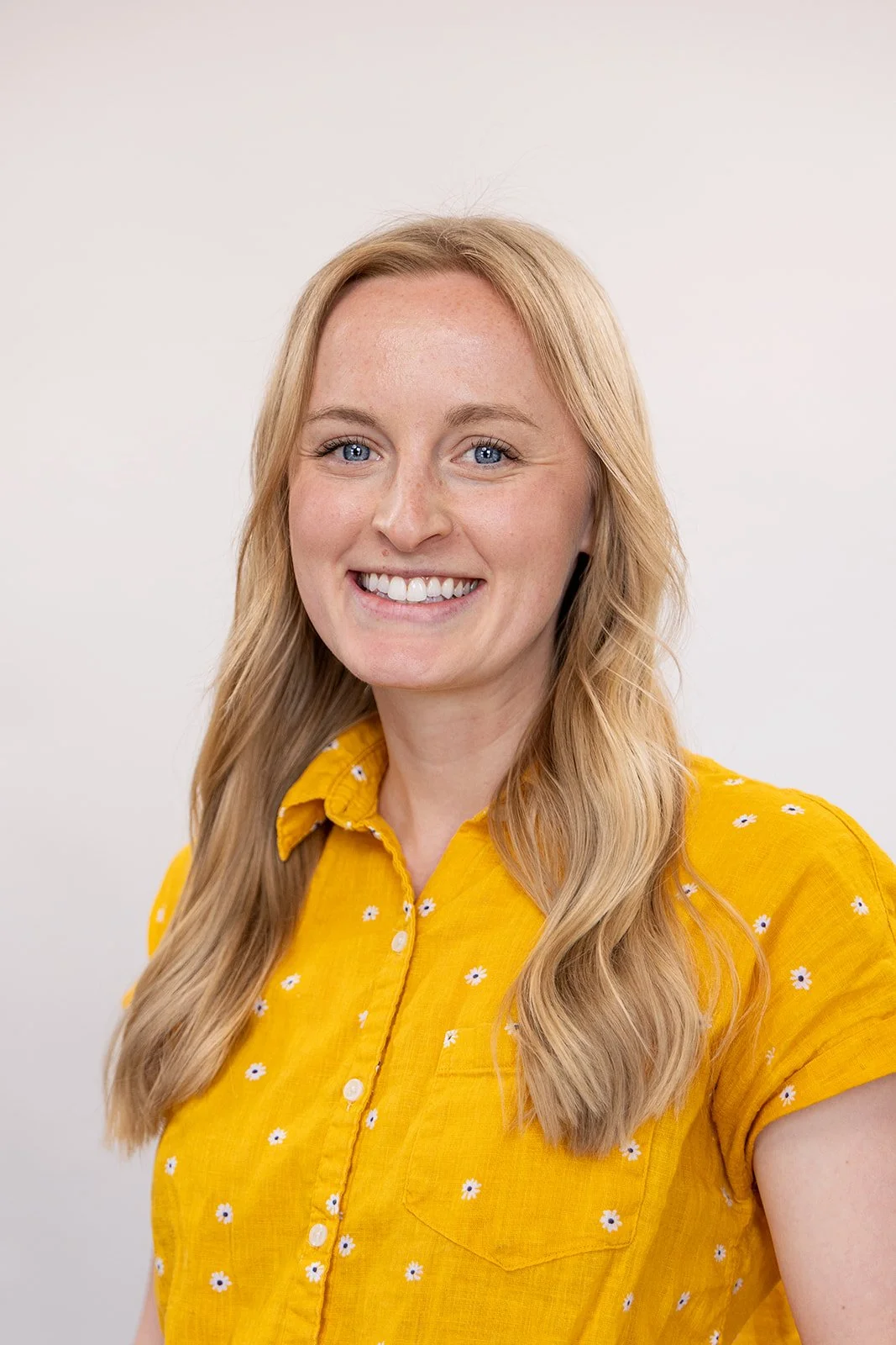 A woman with long blonde hair smiling, wearing a yellow shirt with small white flowers, against a plain light background.