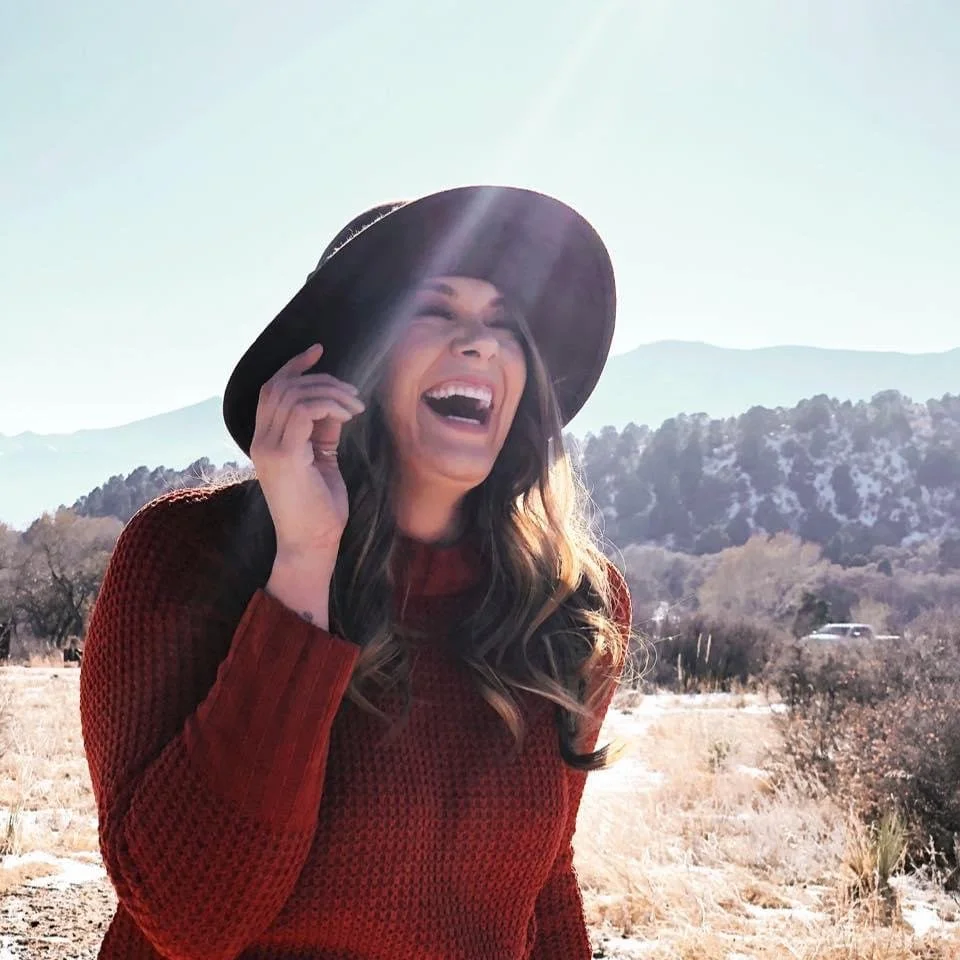 A woman in a red sweater and black hat laughing outdoors in a sunny, mountainous landscape.