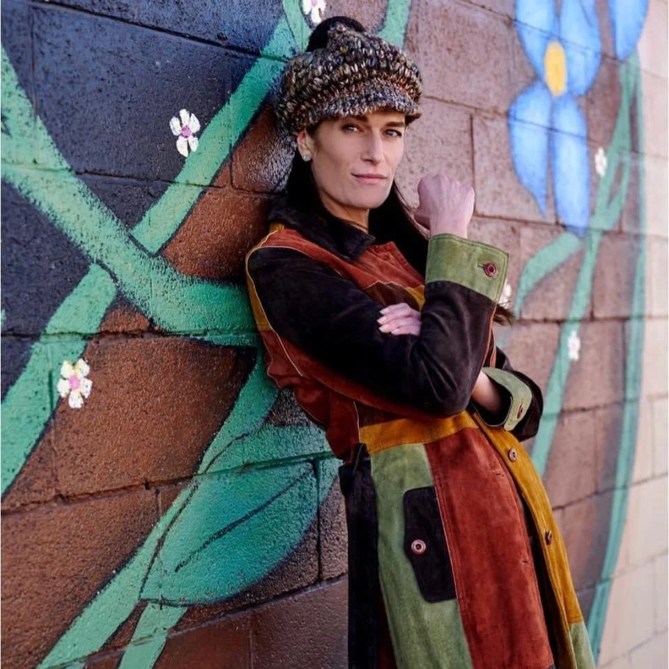 A woman leaning against a brick wall painted with a colorful floral mural, wearing a multicolored coat and a knit hat, making a fist near her face.