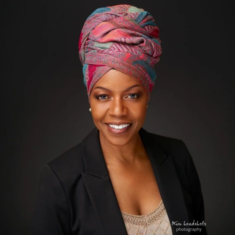 A woman with dark skin, wearing a vibrant multicolored headwrap and a black blazer, smiling against a dark background.