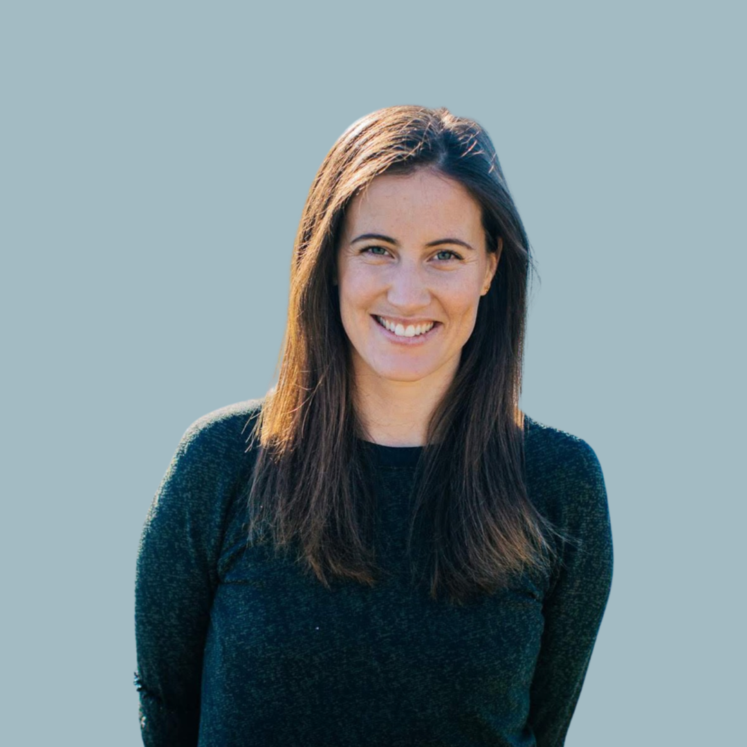 A woman with long brown hair smiling, wearing a black sweater, standing against a plain light blue background. Physiotherapist
