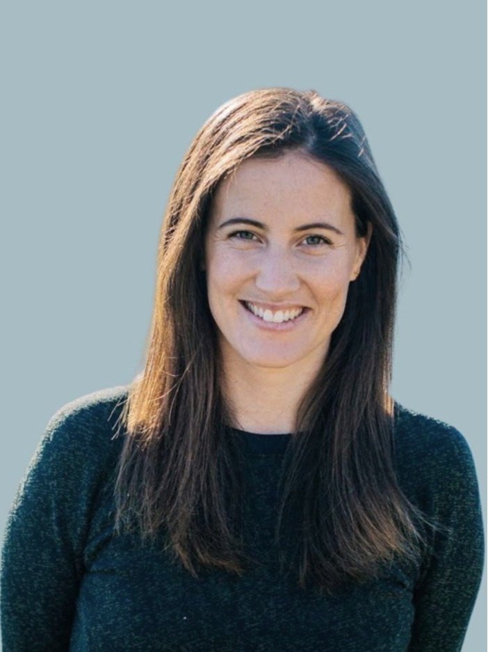 A young woman with long brown hair smiling at the camera against a light gray background.