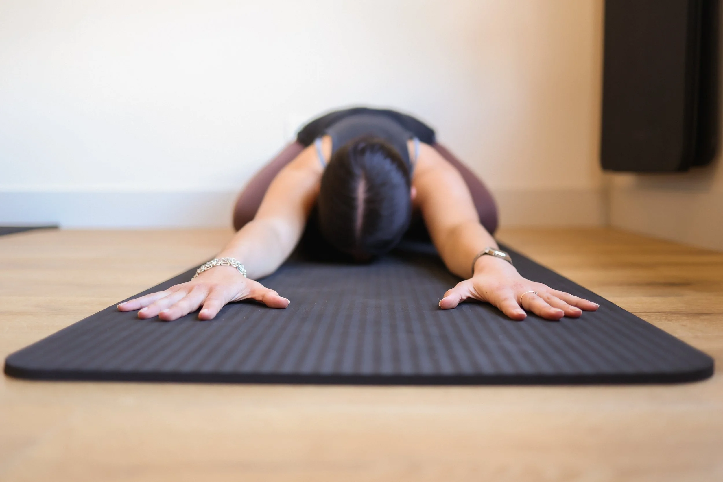 A woman practicing yoga in a low lunge pose on a black yoga mat in a room with light-colored walls.