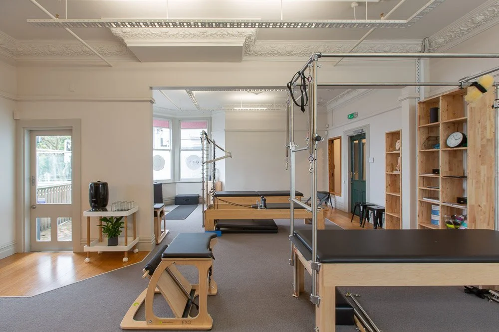 Interior of a Pilates or physical therapy studio with reformer machines, wooden shelves, a clock, and seating area near windows.