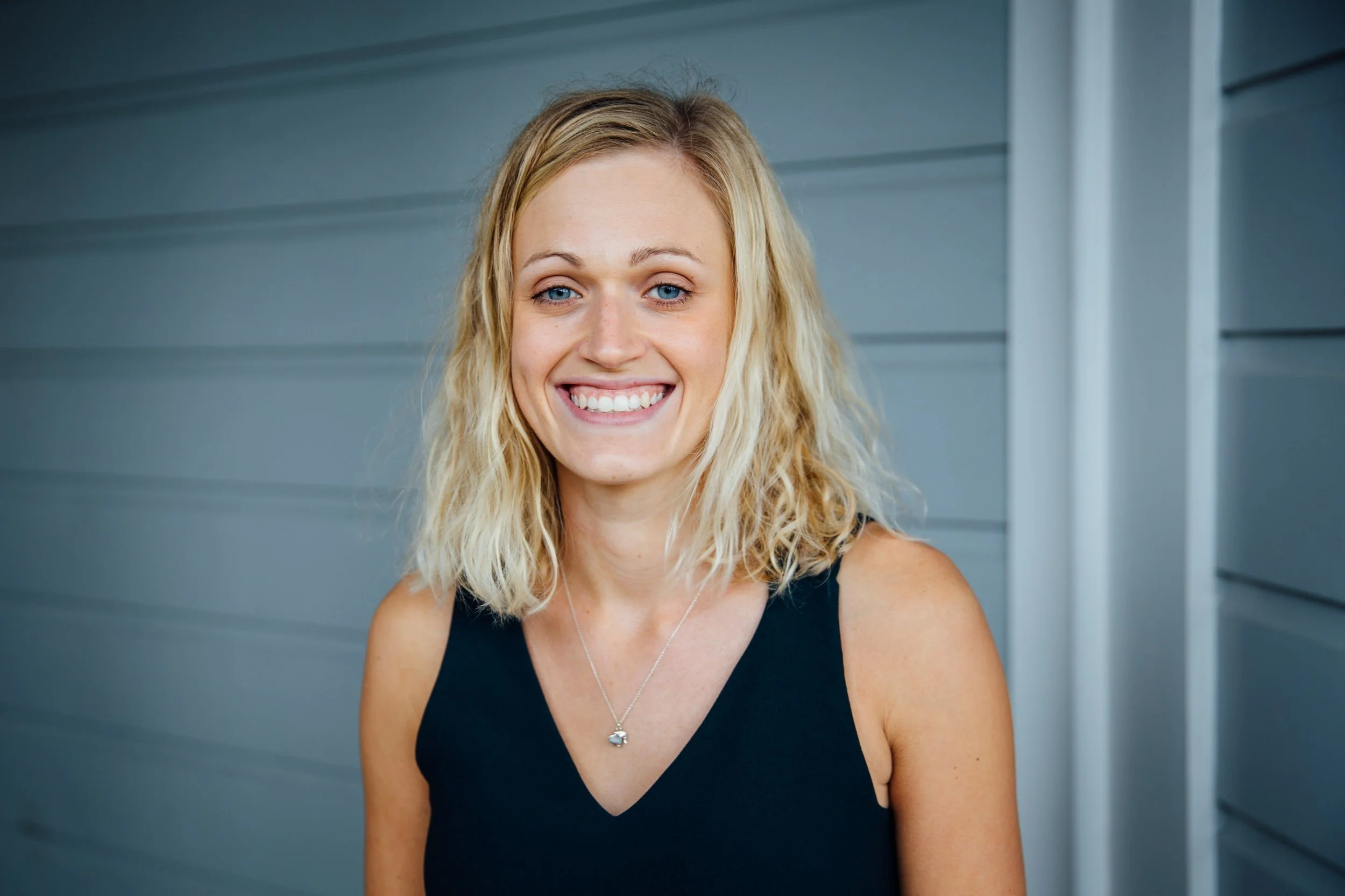 A woman with shoulder-length blond wavy hair smiling in front of a gray horizontal siding wall, wearing a black sleeveless top and a small silver necklace with a charm.