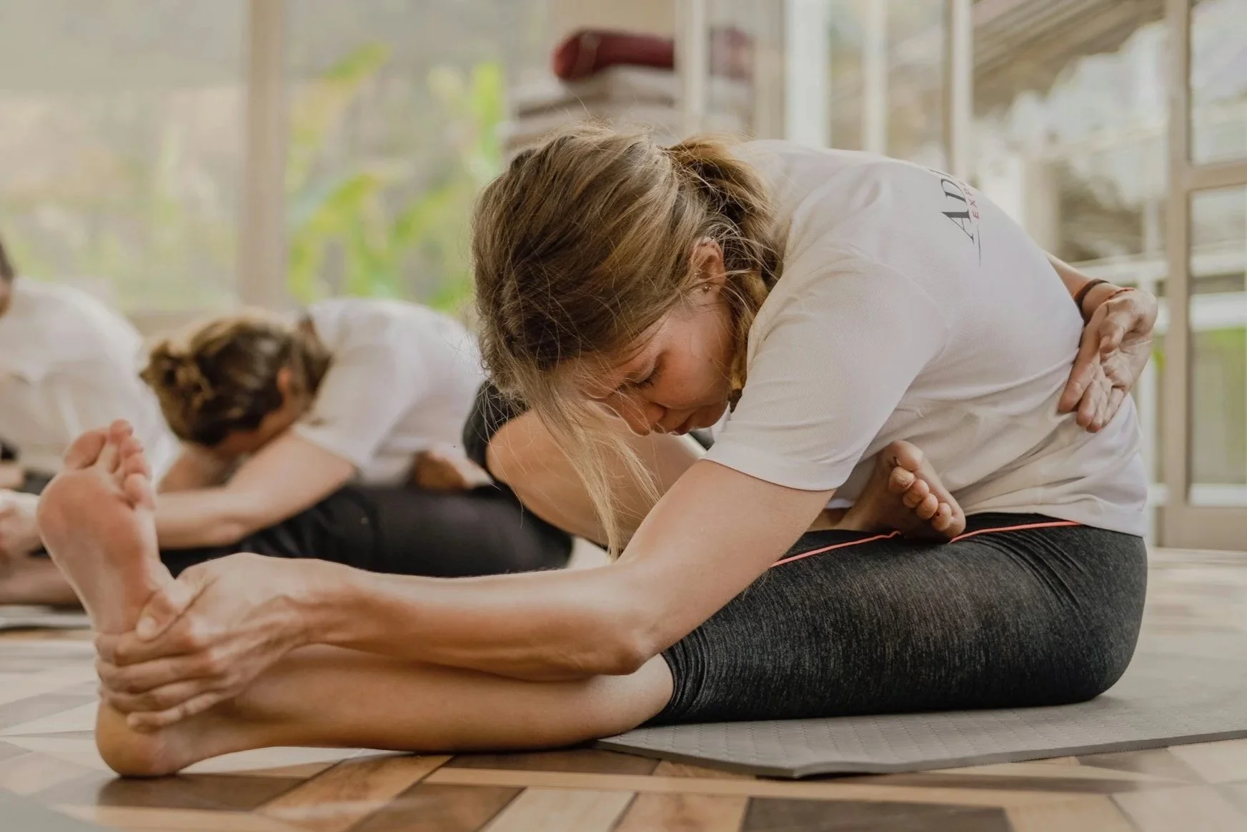 People participating in a yoga class doing seated forward bend stretch on yoga mats inside a bright room.