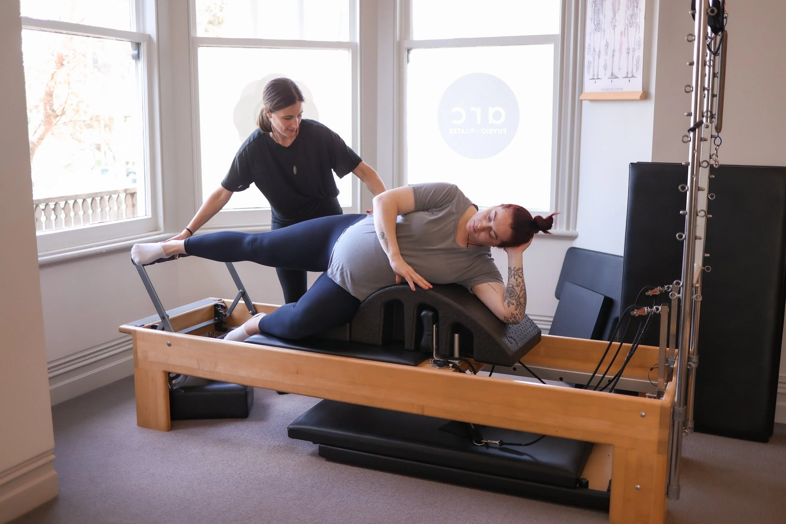 A woman performing physical therapy exercises with a therapist in a room with large windows, a black chair, and posters on the wall. Pelvic Health, Womens health Physio Pilates.