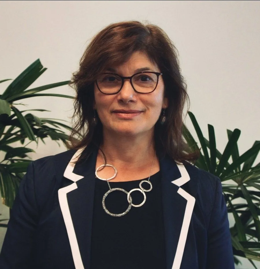 Woman wearing glasses and a necklace with circular pendants, standing in front of plants, wearing a navy blazer with white trim.