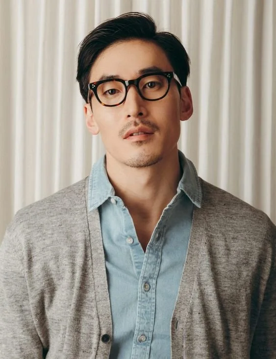 A young man with black hair, glasses, and a mustache, wearing a light blue shirt and gray cardigan, standing in front of a beige curtain.