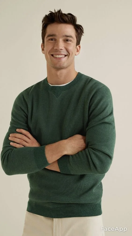A young man with brown hair, smiling, wearing a green sweater with arms crossed, standing against a plain light background.