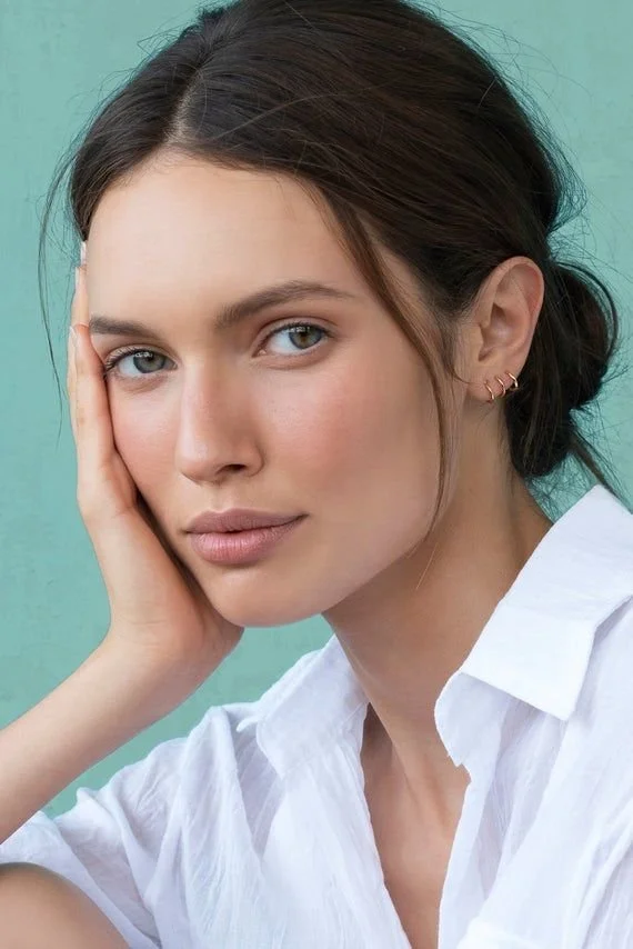 Close-up portrait of a young woman with brown hair, blue eyes, and minimal makeup, wearing a white shirt, touching her face with her hand against a teal background.