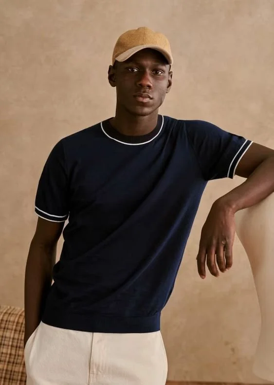Young man wearing a beige cap, navy blue T-shirt, and light-colored pants posing indoors against a neutral background.