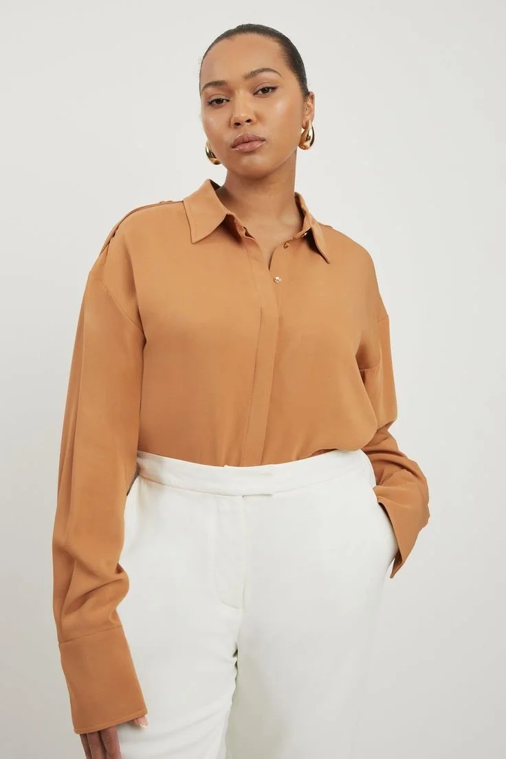 Woman wearing a tan button-up shirt and white high-waisted pants with her hand in her pocket, standing against a plain background.