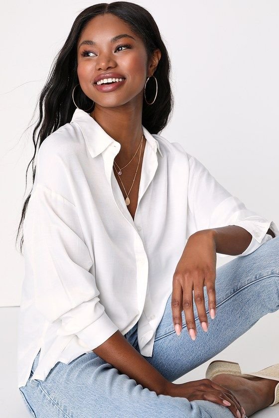A young woman with long black hair wearing a white button-up shirt, light wash jeans, and jewelry, smiling while sitting on the floor against a plain white background.