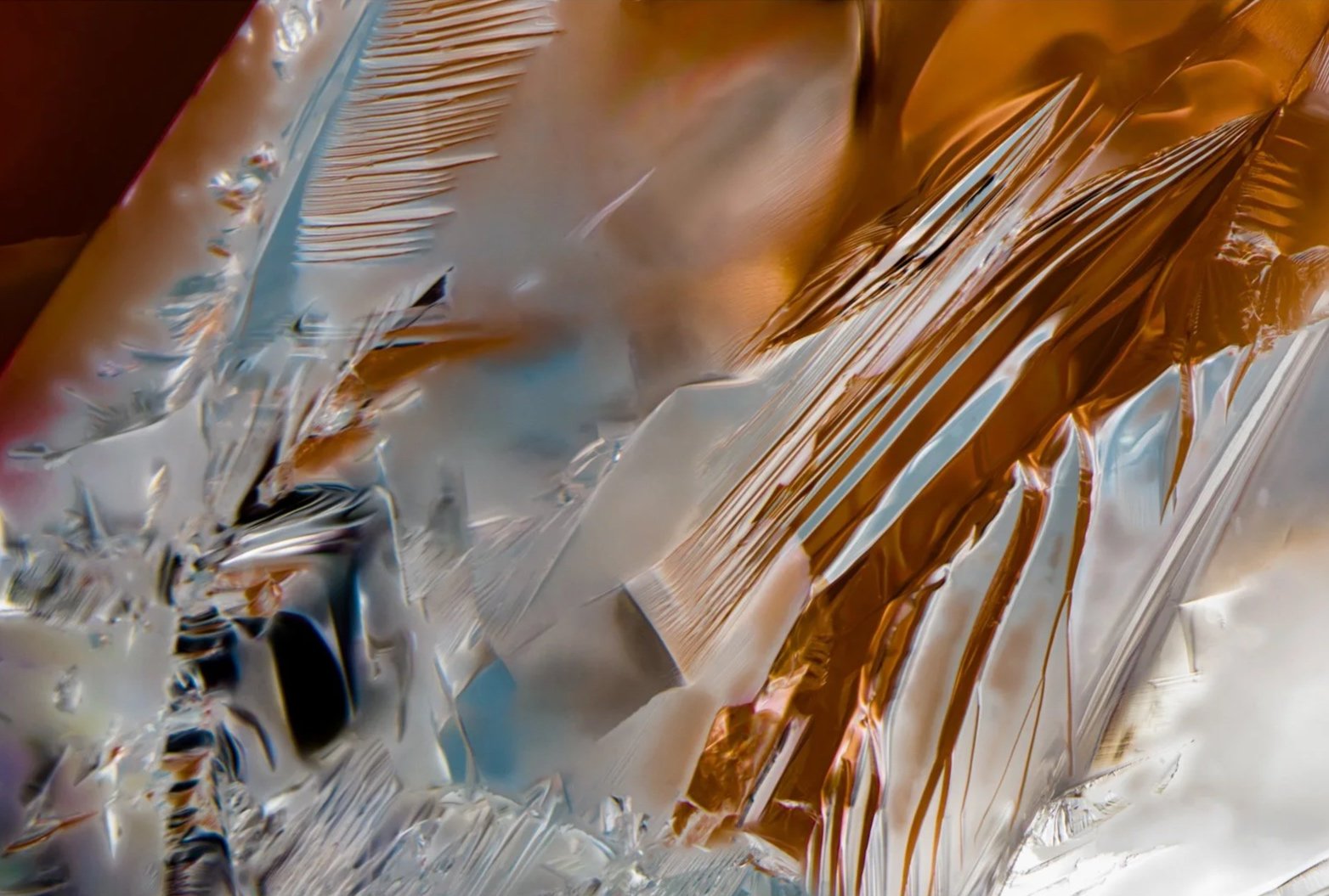 Macro photograph by Cesar Cueva revealing layered crystalline structures, fine parallel fractures and translucent planes in amber, silver and pale blue.