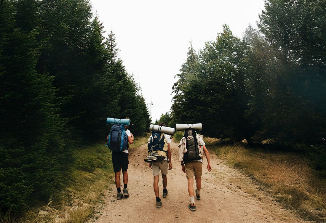 Trois randonneurs marchant sur un chemin forestier avec des sacs à dos et des tapis de yoga roulés sur leurs épaules, entourés d'arbres vert foncé.