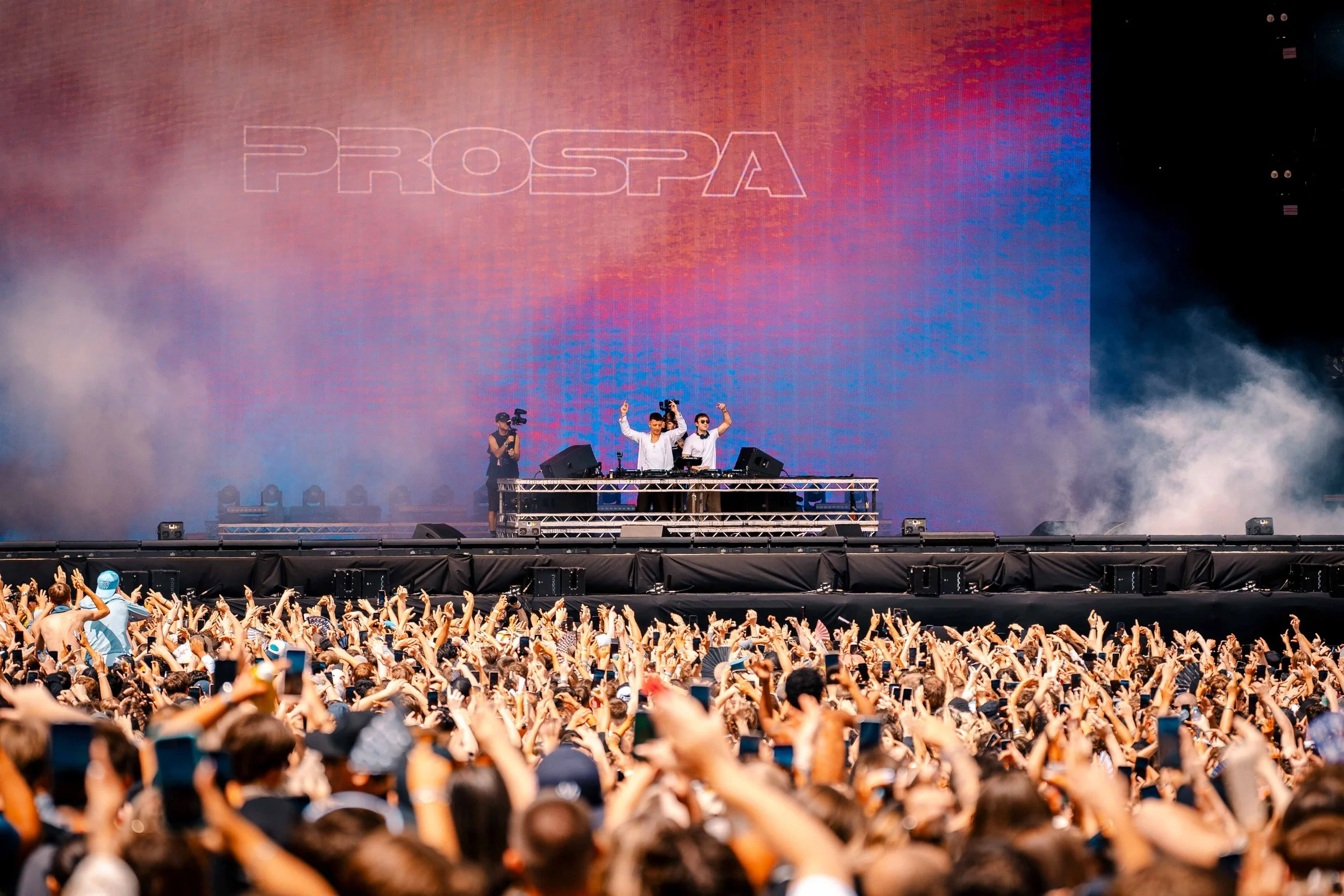 A large outdoor concert with a crowd of people and a stage where a DJ performs. The crowd has raised hands and some are taking photos. The stage has a large digital screen displaying the word 'PROSPA' and fog effects.