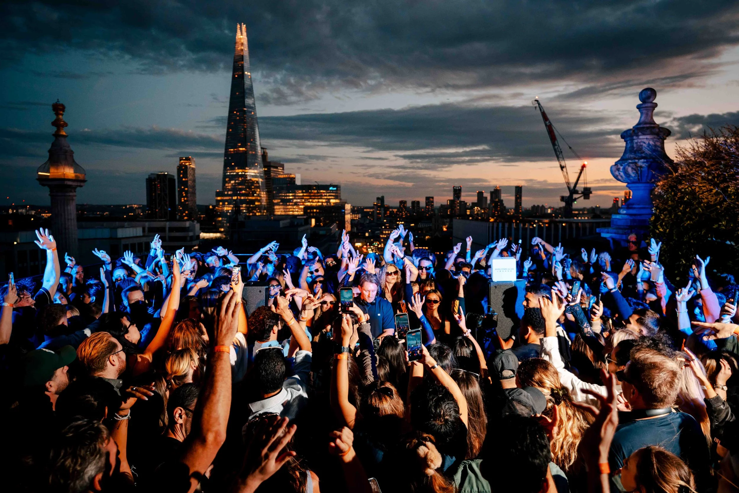 A large crowd of people gathered outdoors at sunset, dancing and taking photos with smartphones, with city skyscrapers, including the Shard, and construction cranes in the background.