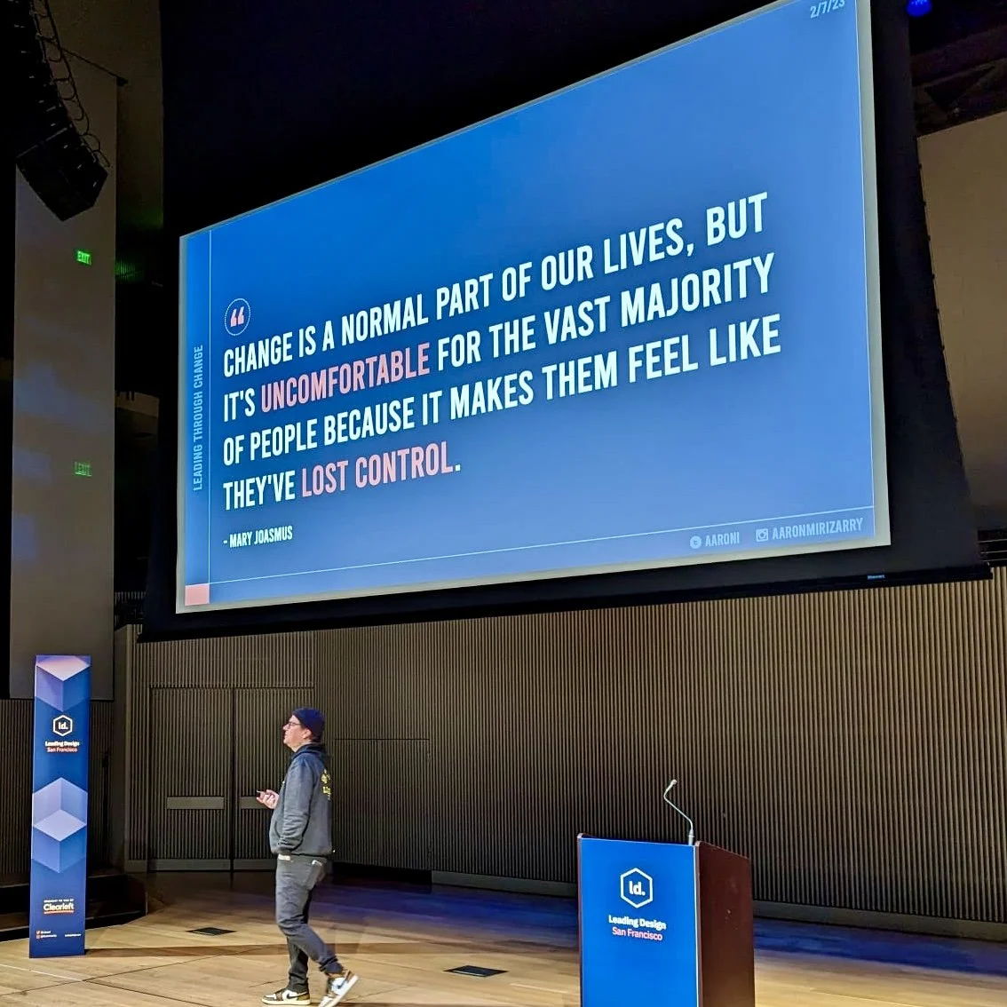 A large screen displaying a quote about change, with a man walking in front of it inside a conference or presentation room. The room has a wooden floor and a podium with a microphone. The quote says: "Change is a normal part of our lives, but it's uncomfortable for the vast majority of people because it makes them feel like they've lost control" by Mary Joamus. There are signs and branding for ID Leading Design, San Francisco.