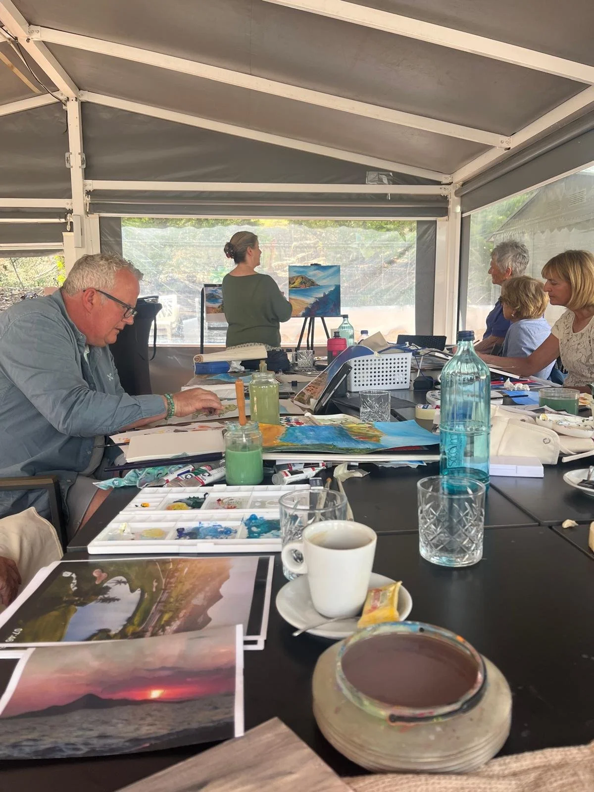 A group of people sitting around a table in a bright tent, engaged in a painting class, with supplies, paintings, a coffee cup, and water bottles on the table, and an instructor demonstrating landscape painting at the front.