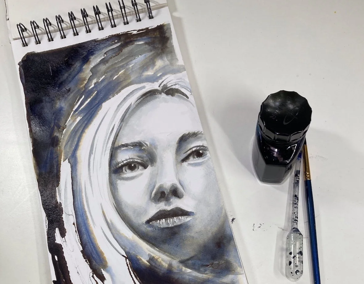 A watercolor portrait of a woman's face on a spiral-bound sketchbook page, with paint bottles and brushes beside it.