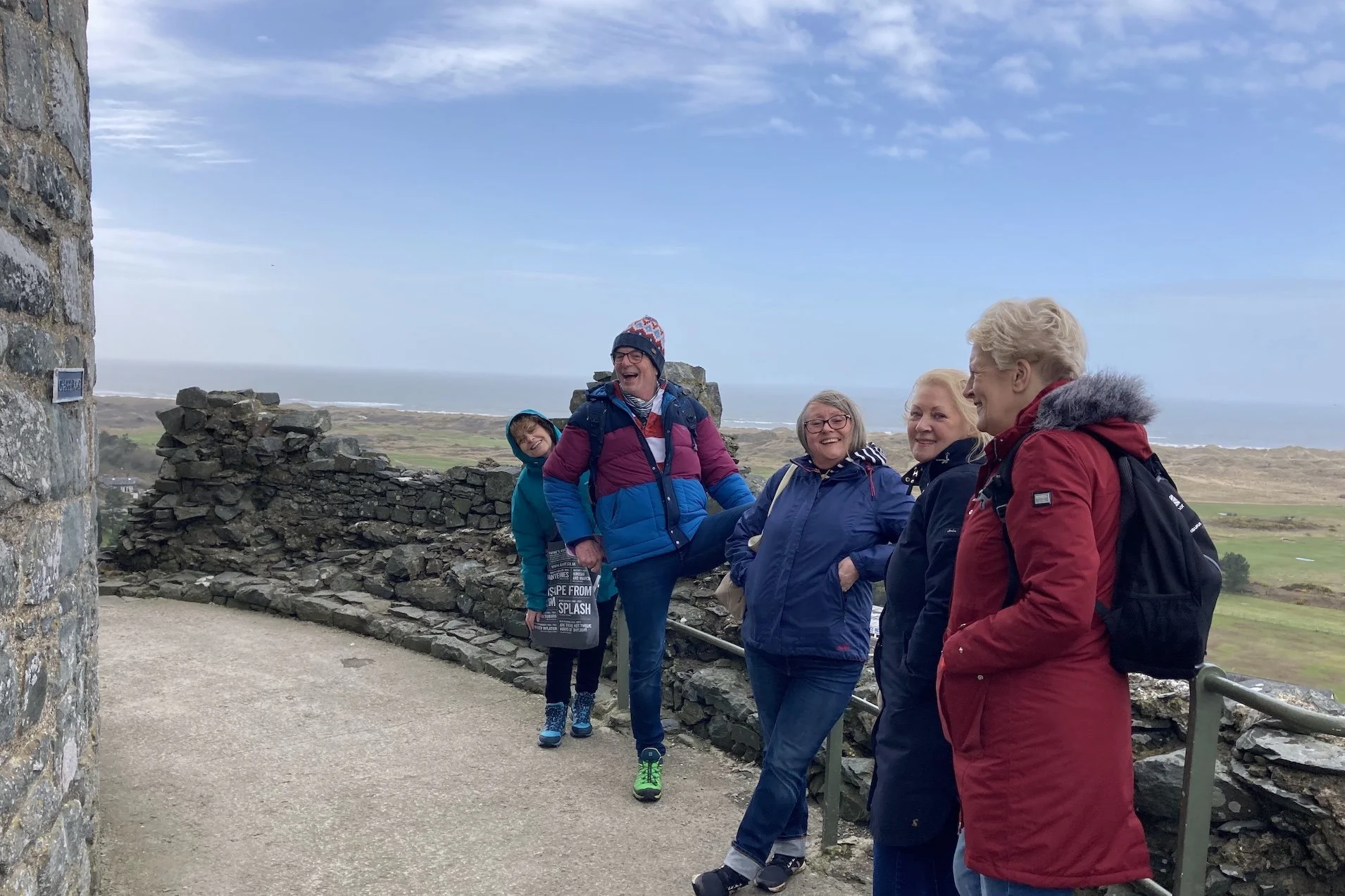 A group of five people smiling and enjoying a scenic view near a stone wall, with a cloudy sky and ocean in the background.