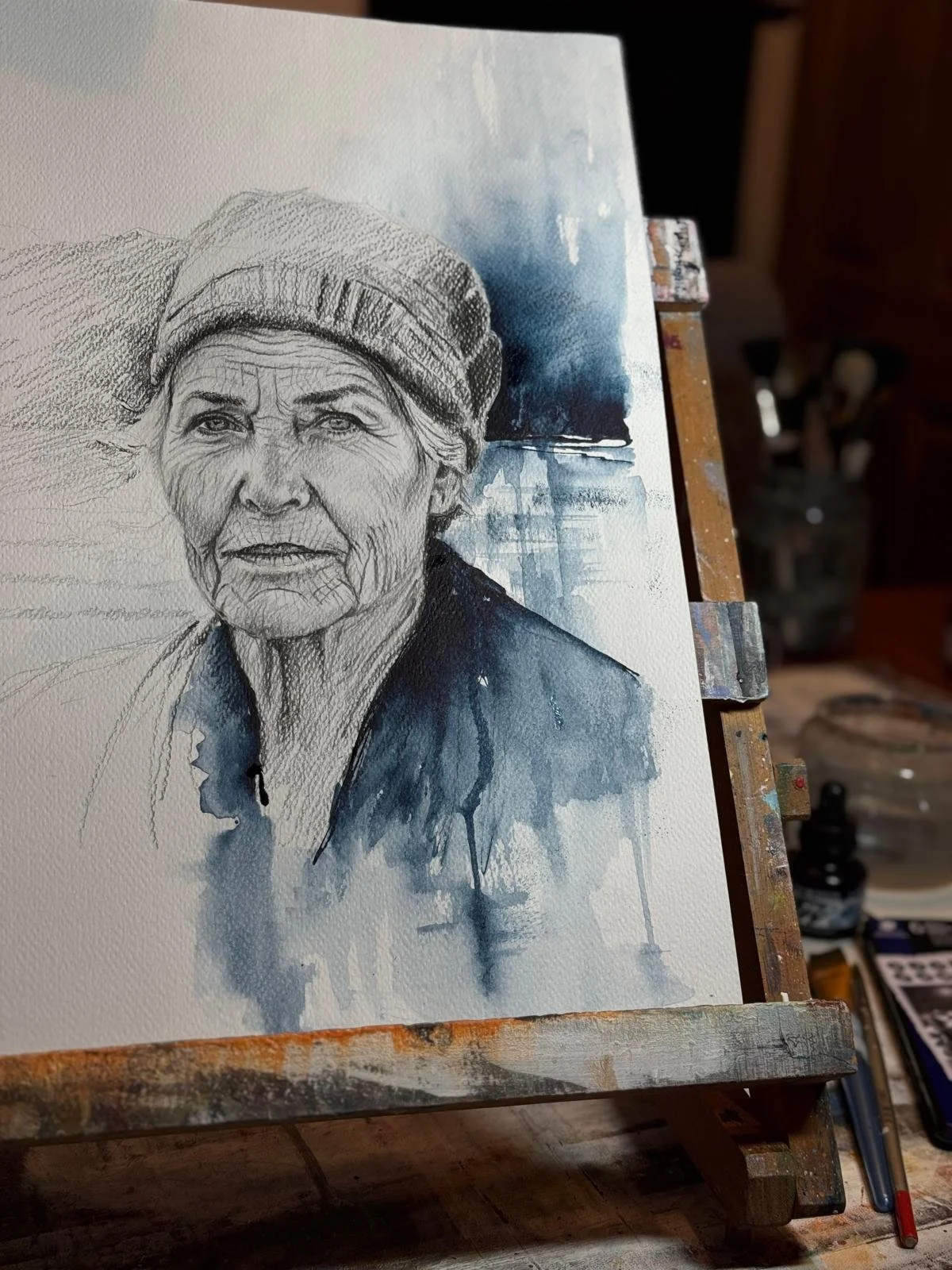 A watercolor painting of an elderly woman wearing a knit hat and dark clothing, with a not yet finished background.