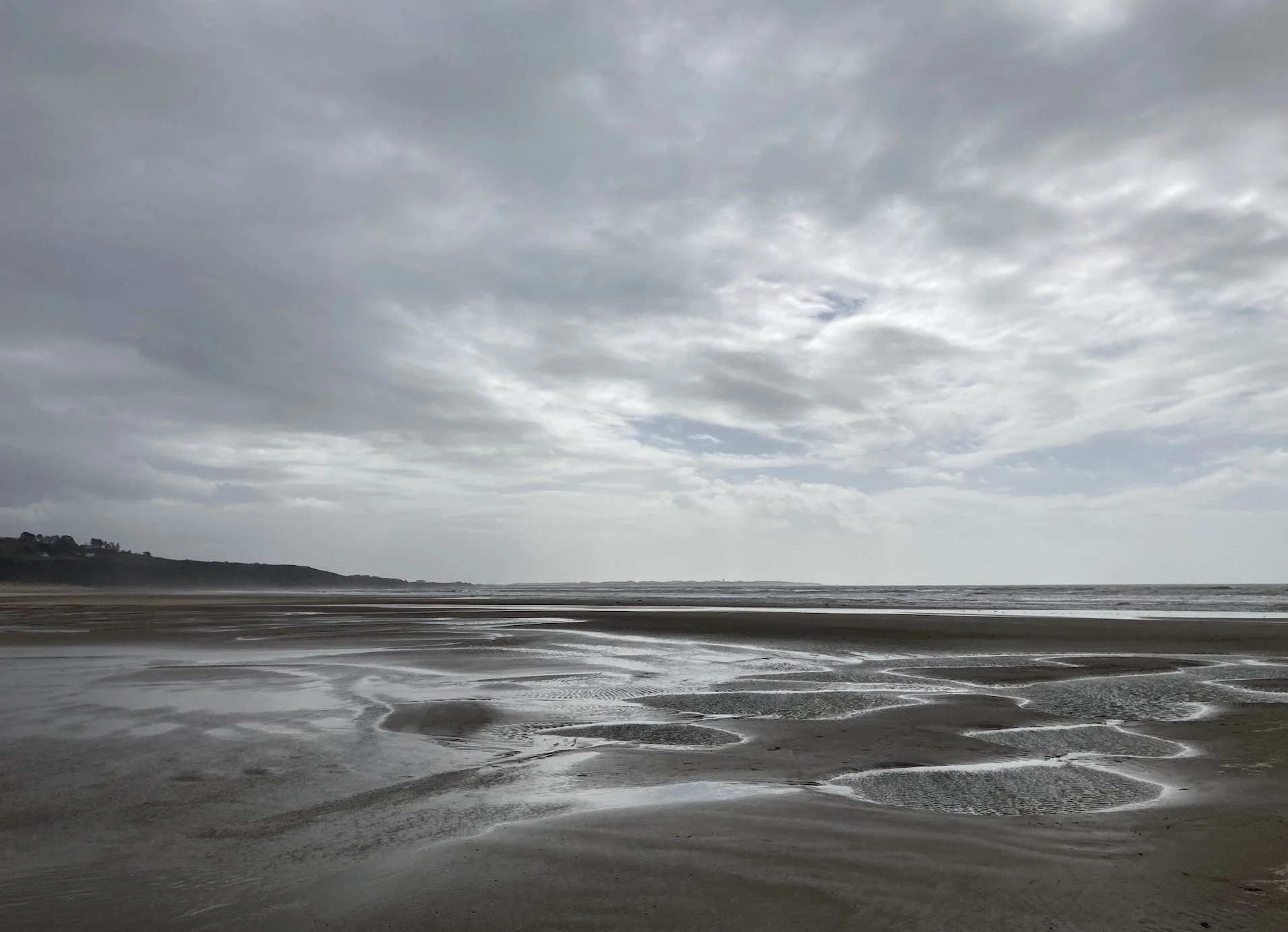 Overcast beach with tidal pools and wet sand, distant shoreline and cloudy sky.