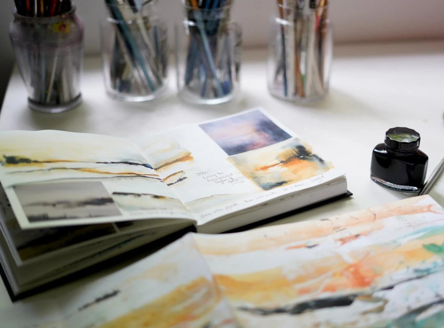 Open sketchbook with watercolor paintings, three glass jars filled with paintbrushes, a bottle of black ink, and watercolor paintings spread out on a white table.