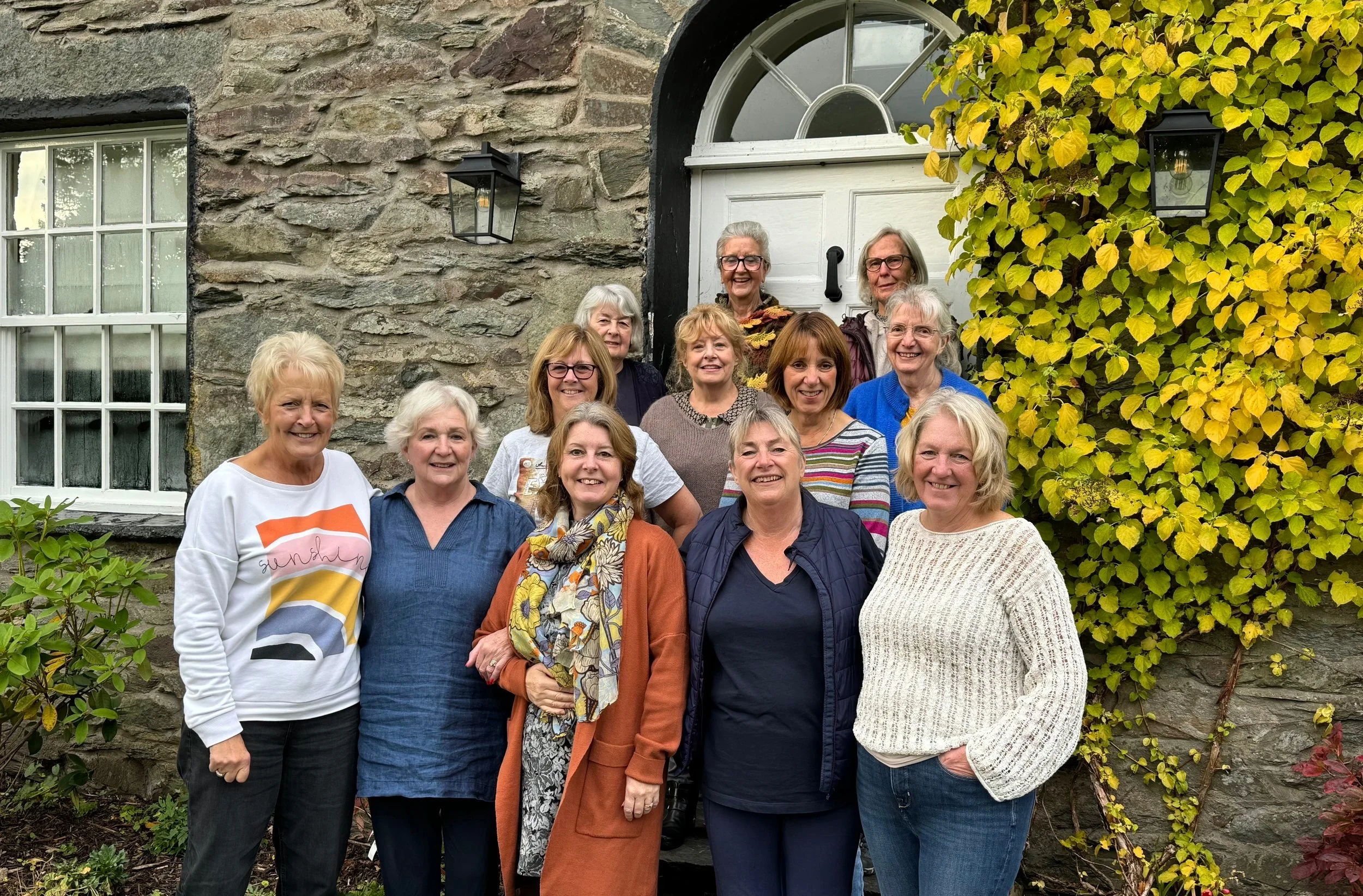 Group of ten senior women standing in front of a stone house with a white door and yellow-green ivy, smiling outdoors.