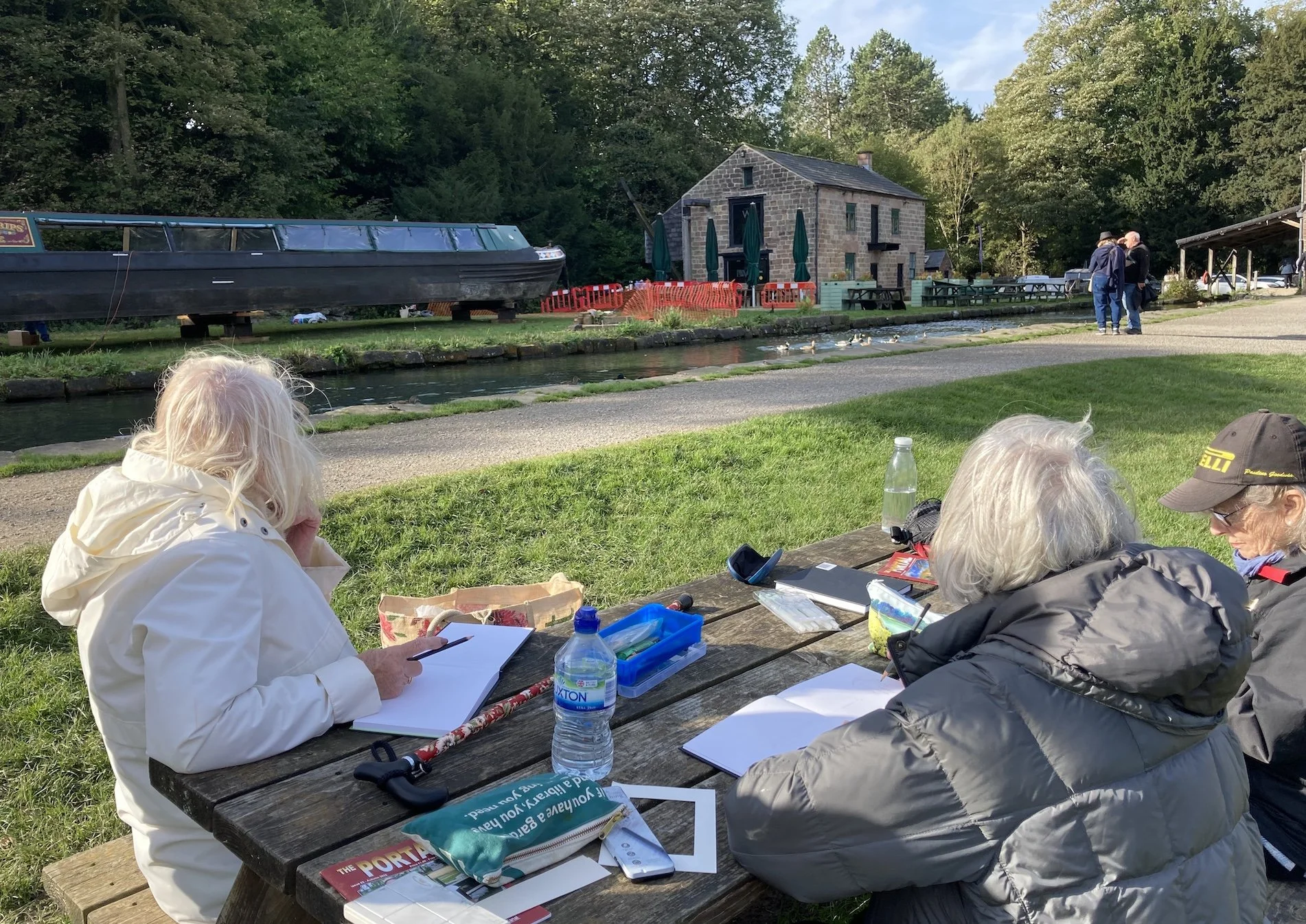 Three elderly women sitting at a wooden picnic table outdoors near a small canal, observing ducks, with trees, a stone building, and a boat in the background.