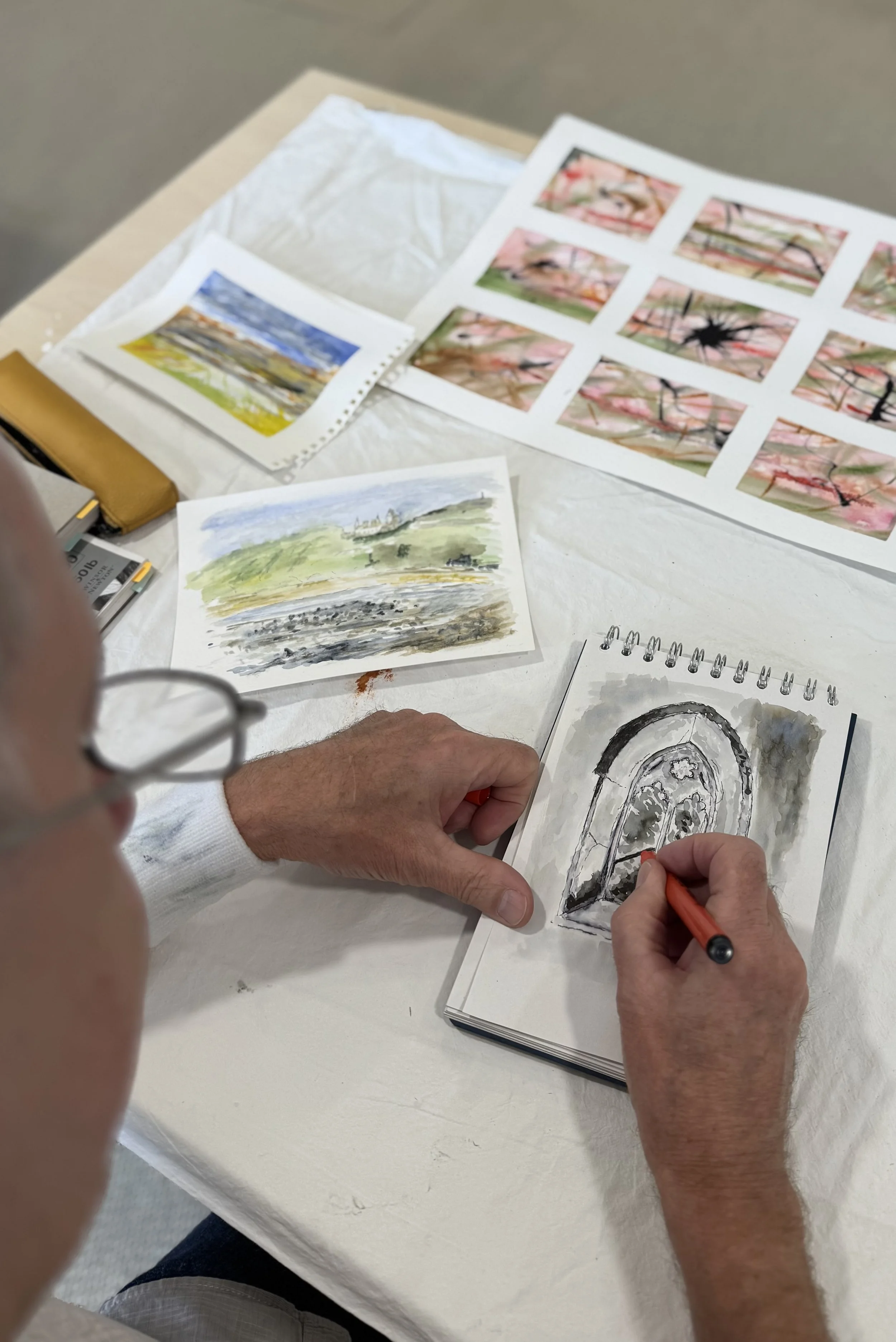 An artist is drawing a black and white ink illustration of an arched door or window in a sketchbook at a table with watercolor paintings and printouts of landscape and floral images.