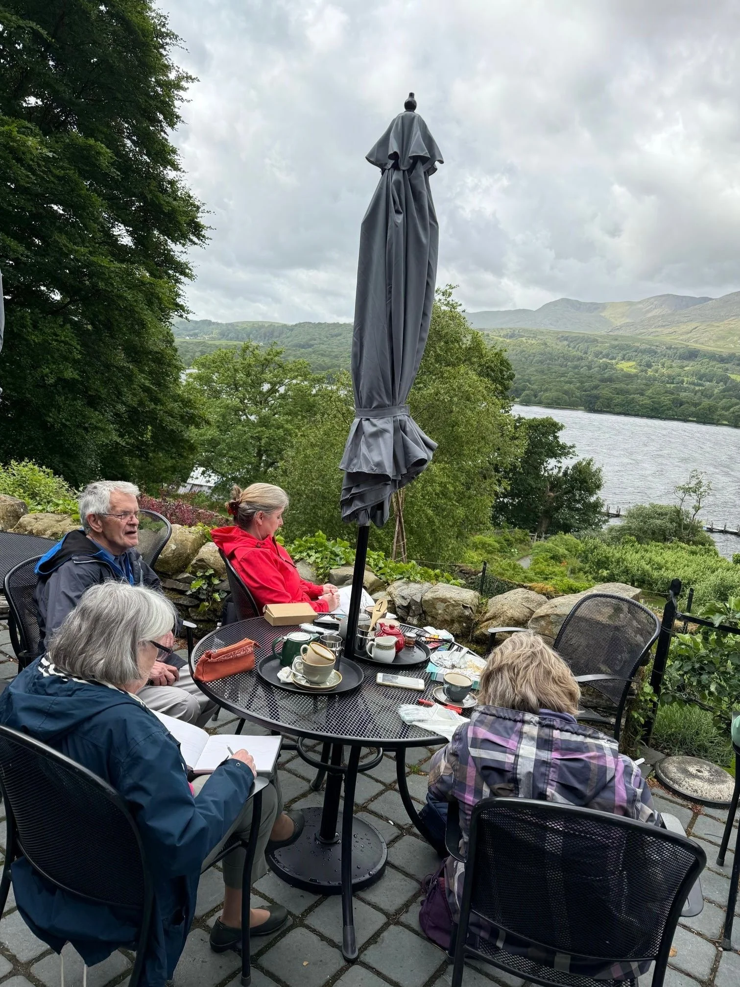 Four people sitting at a round outdoor table with cups, teapots, and notebooks, overlooking a lake and mountains, with a closed gray umbrella in the center.