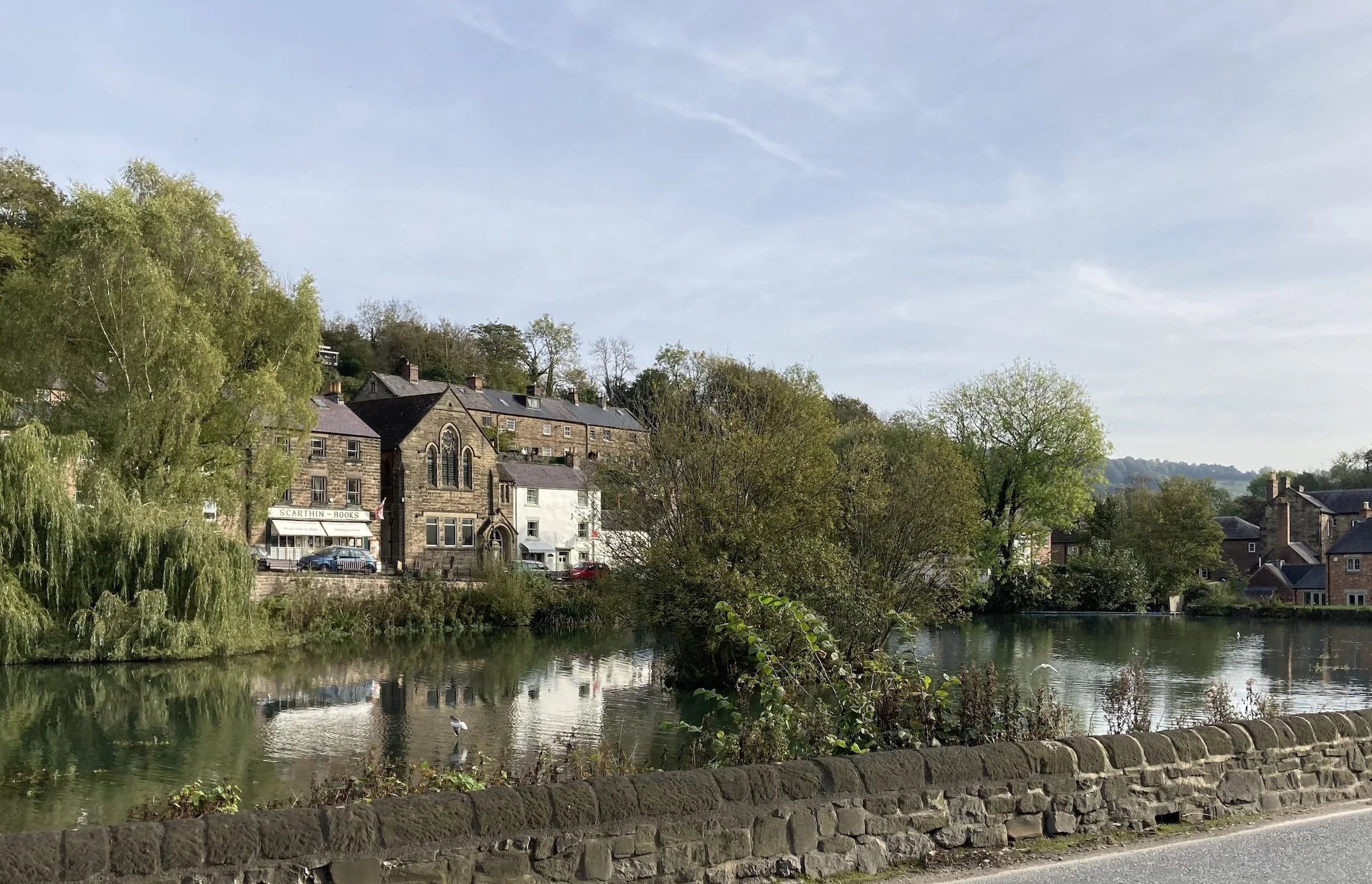 A scenic view of a small town with stone buildings by a calm river, surrounded by green trees, under a partly cloudy sky.