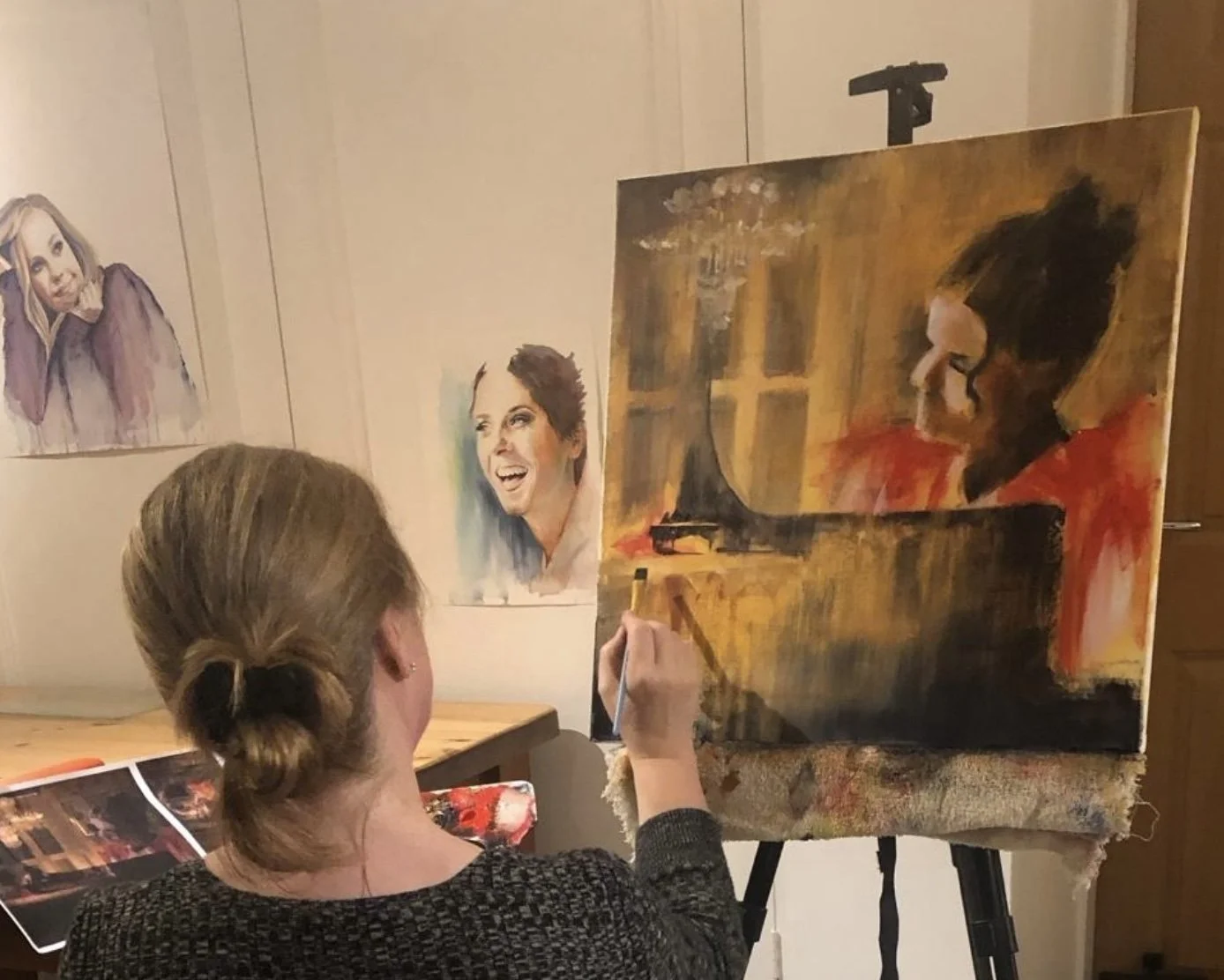 A woman with her hair in a bun painting an abstract portrait of a woman with a dark bun and red top on a canvas. The room has several other portraits on the wall, including a woman with blonde hair and a smiling woman with short hair.