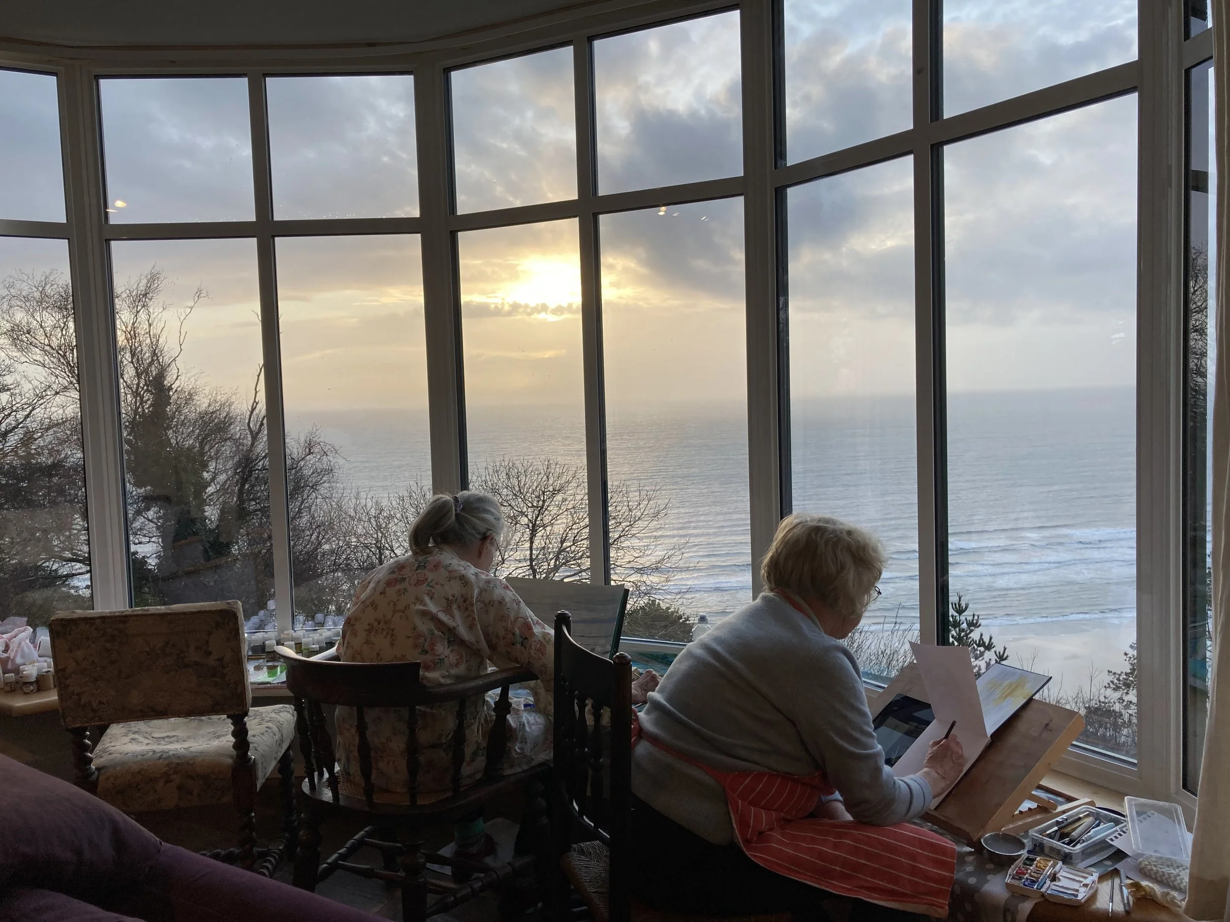 Two women sitting in a sunlit room with large glass windows overlooking a beach and sunset, engaged in art or craft projects.