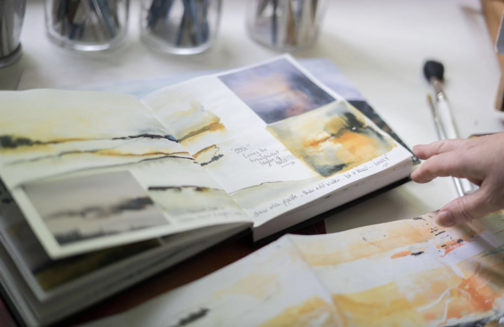 Open sketchbook with watercolor paintings of landscapes on a desk, surrounded by paint jars and brushes.