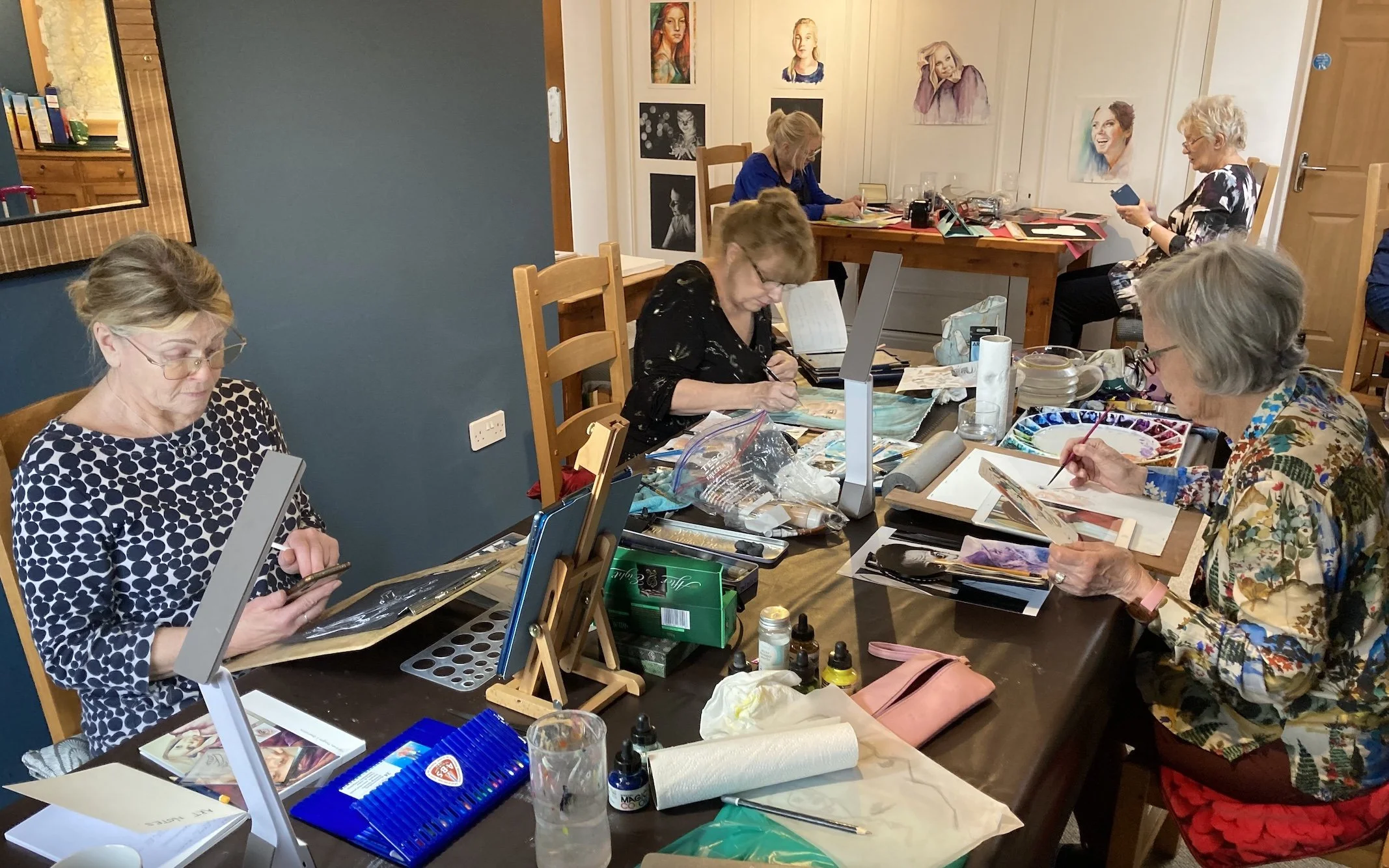 A group of women, mostly seniors, are sitting around a large table engaged in art projects, including watercolor painting and sketching, with art supplies scattered across the table.