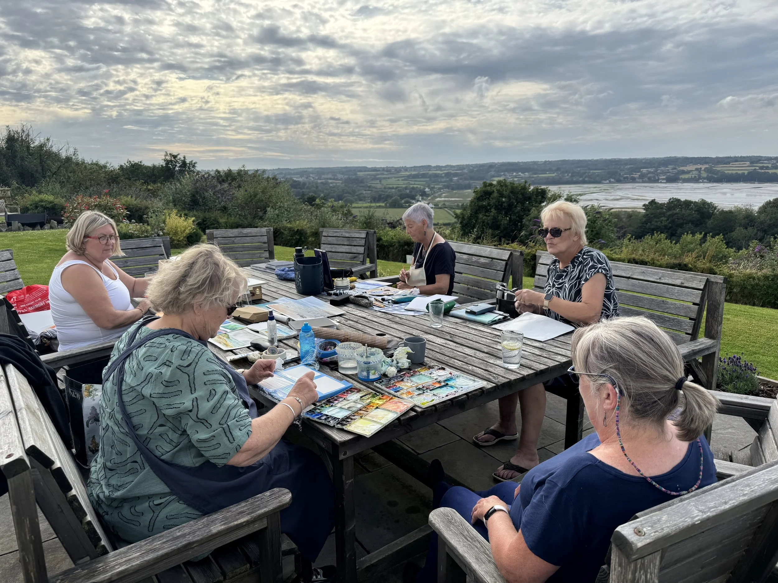 Six women sitting around an outdoor table on a patio, engaging in arts and crafts while overlooking a scenic landscape with fields and water.