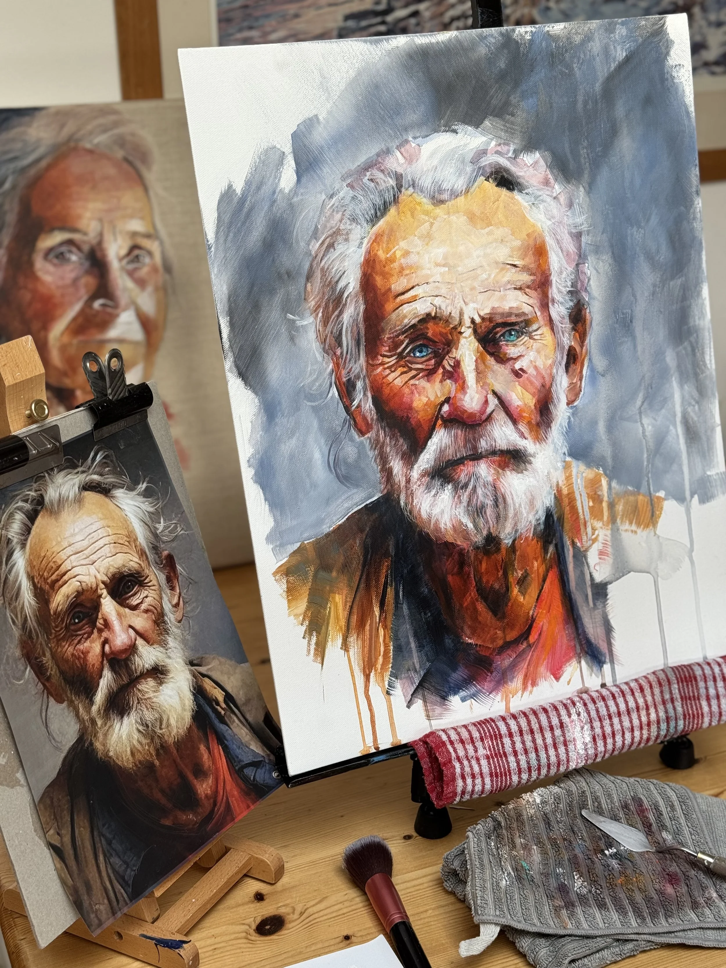 An artist's painting of an elderly man with white hair and beard, wearing a dark shirt, with a background of blue and gray tones, on an easel. There is also a reference photo of the same man on a smaller easel nearby, showing a man with similar white hair, beard, and a weathered face. The workspace has brushes, a knife, and a cloth with paint on it on a wooden table.