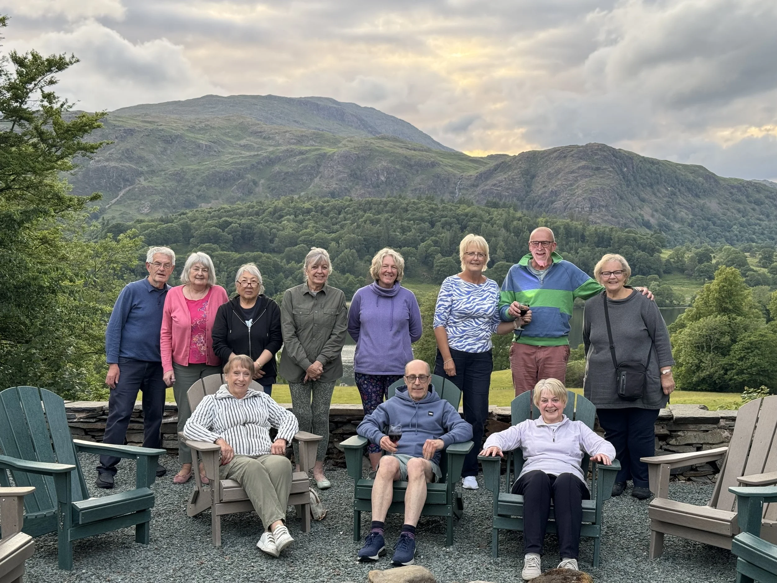 A group of eleven people, mostly seniors, posing outdoors with a scenic mountain view in the background. Some are sitting in chairs, others standing, holding glasses of wine, on a gravel surface with outdoor furniture around.