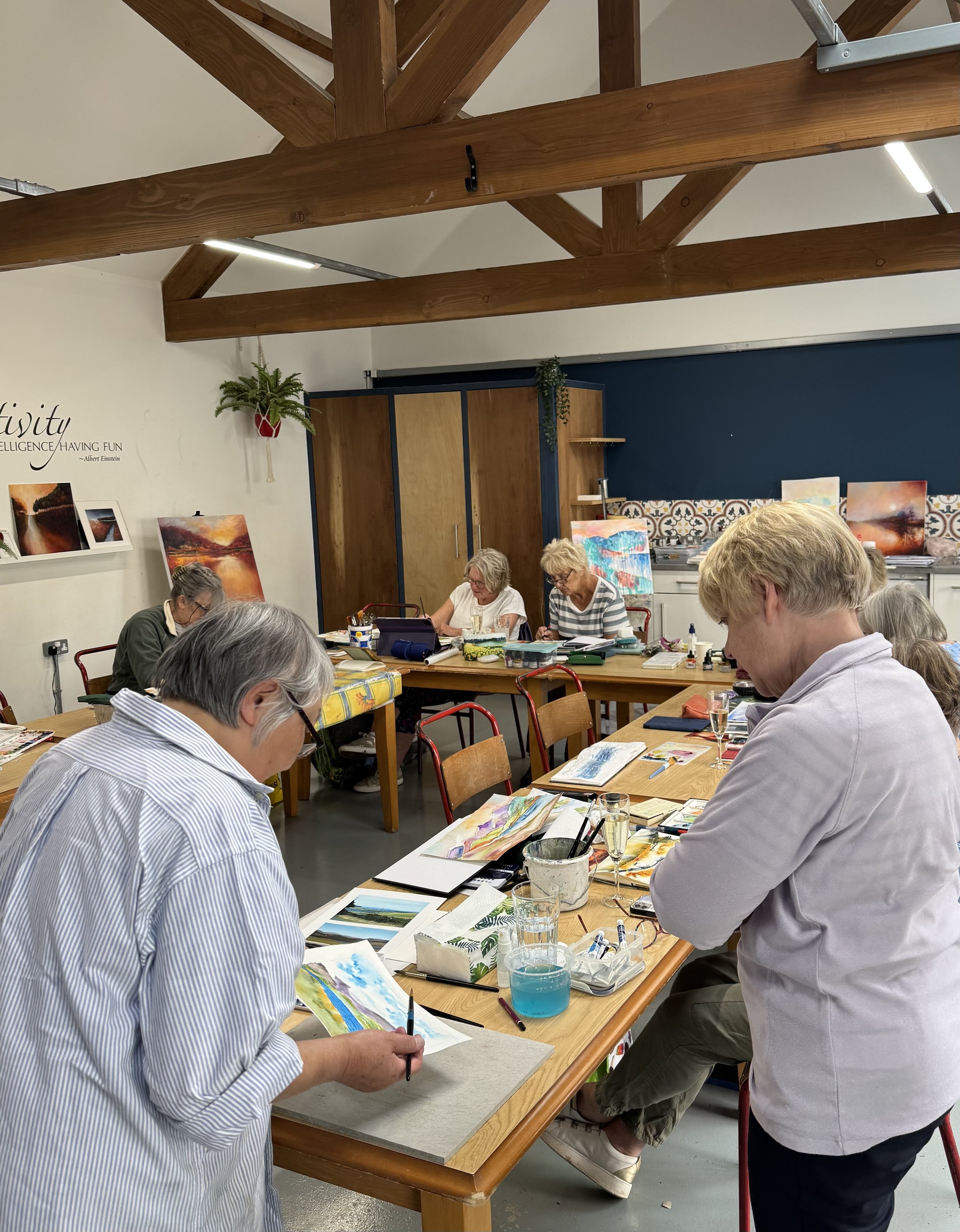 A group of older adults painting watercolors at a table in an art studio, with several completed paintings on display and shelves on the wall.