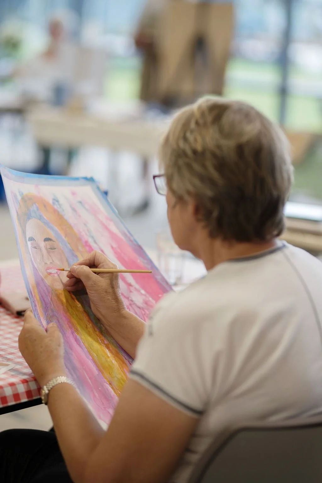 An older woman with glasses and a wrist bracelet is painting a colorful portrait of a person with blonde hair on a canvas at a table with a red and white checkered tablecloth in a bright room with large windows.