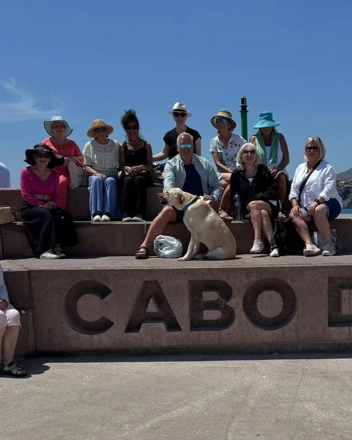 Group of people sitting on a stone ledge with "CABO" engraved in front, on a sunny day with a clear blue sky, some wearing sunglasses and hats, with a dog sitting near the center.